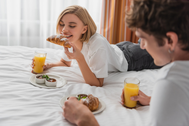 happy woman with orange juice eating tasty croissant near blurred boyfriend on bed in hotel apartments - Photo, Image