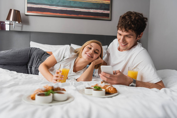 молодой человек пользуется мобильным телефоном рядом с улыбающейся подругой и завтракает на кровати в отеле - Фото, изображение
