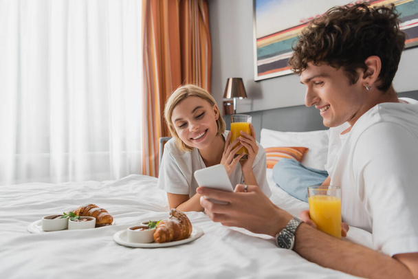 молодой человек показывает мобильный телефон улыбающейся девушке рядом с круассанами и апельсиновым соком на кровати отеля - Фото, изображение
