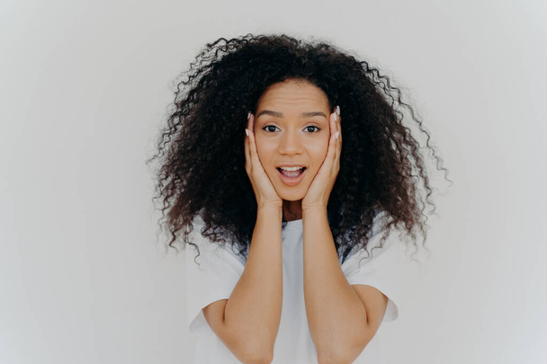 Photo of surprised cheerful woman with Afro haircut, keeps both hands on cheeks, has natural beauty, opens mouth, cannot believe in exciting news, wears t shirt, poses against white background. - Photo, Image