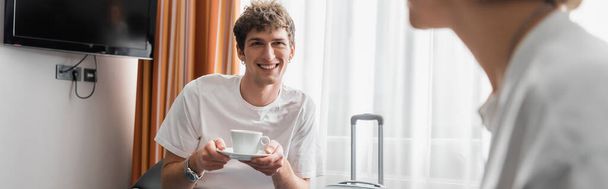happy young man holding coffee cup near girlfriend in hotel room on blurred foreground, banner - Photo, image