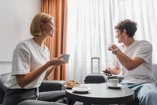 young man holding smartphone and pointing at window near girlfriend with coffee cup in hotel room - Фото, зображення