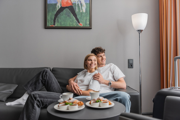 весела пара обіймається на дивані біля смачних круасанів і чашок кави в готелі
  - Фото, зображення
