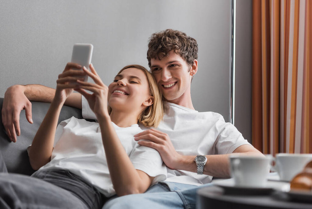 веселая блондинка с мобильным телефоном рядом с молодым парнем и размытые кофейные чашки на диване в отеле - Фото, изображение