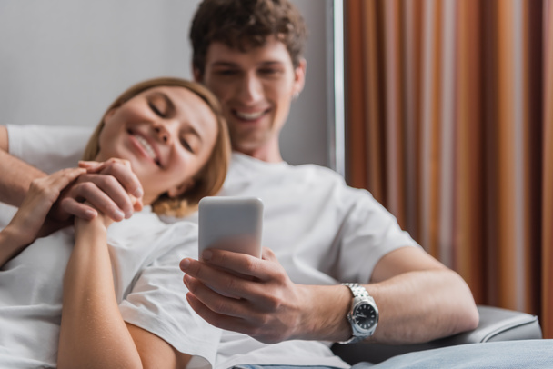young and happy man holding cellphone while resting and holding hands with girlfriend on blurred background in hotel room - Foto, Imagen