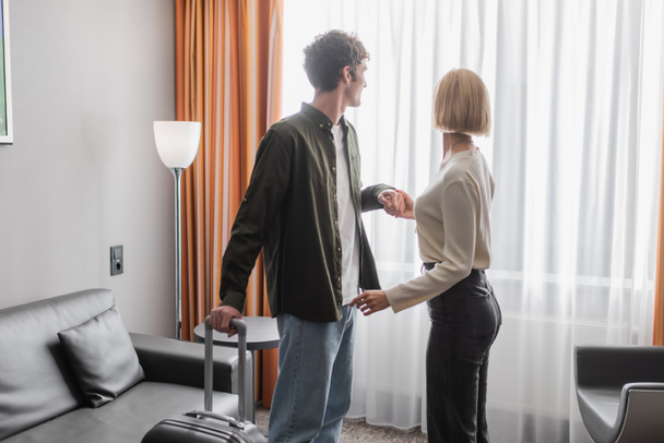 couple of young tourists with travel bag holding hands near window with curtains in hotel suite - Foto, imagen