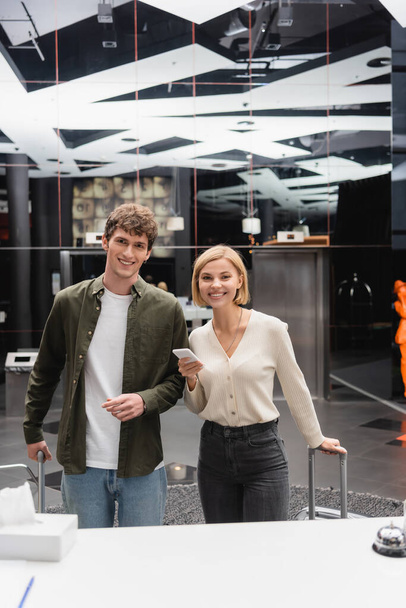 young man and cheerful blonde woman with mobile phone looking at camera near reception in modern hotel - Fotoğraf, Görsel