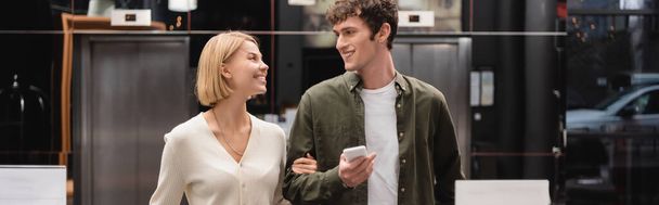 cheerful blonde woman and man with mobile phone looking at each other in hotel lobby, banner - Foto, immagini