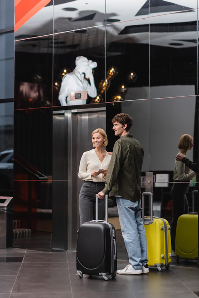 улыбающаяся пара с чемоданами, смотрящими в сторону, стоя с дорожными сумками в современном отеле - Фото, изображение
