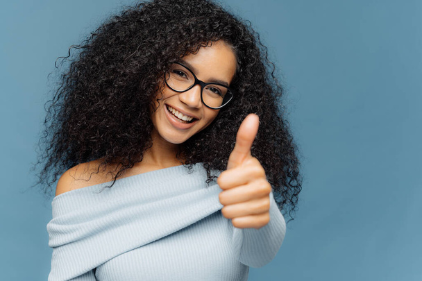 Its excellent! Glad smiling African American woman shows like gesture, keeps thumb up, gives approval, cheers over blue wall, dressed in casual clothes, expresses approval of amazing product - Valokuva, kuva