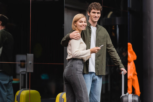 happy young man and blonde woman with mobile phone looking at camera near travel bags in hotel - Foto, Imagen