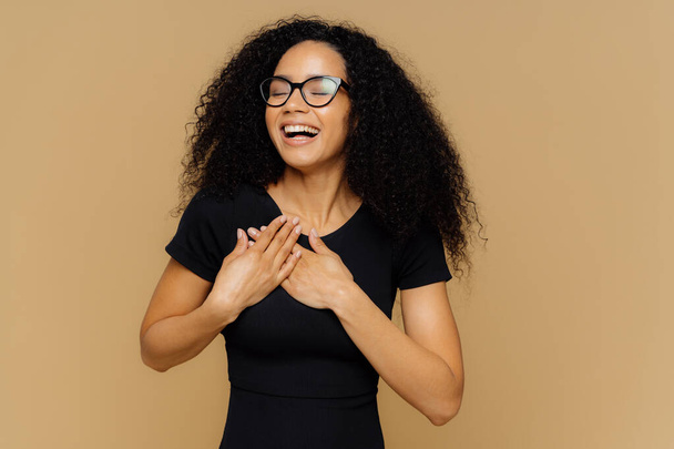 Thankful positive woman smiles happily, makes gratitude gesture, keeps hands on chest, smiles broadly, wears casual t shirt, optical glasses, closes eyes from pleasure, isolated on beige background - Photo, Image