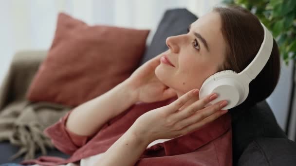 Lähikuva nuori söpö valkoihoinen nainen istuu sohvalla ja kuunnella musiikkia valkoisissa kuulokkeissa.Lady nauttia äänen rentouttava kotona. Ihmiset ja dekhnologia käsite - Materiaali, video