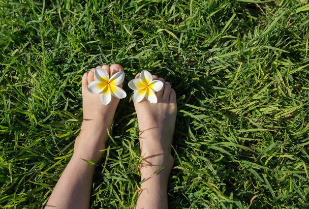 A gyerek mezítláb fekszik egy zöld gyepen, két pluméria virággal a lábujjai között. öröm, vidám pozitív légkör, boldog gyermekkor, pihenés. Helló nyár. a természet energiája. Föld napja - Fotó, kép