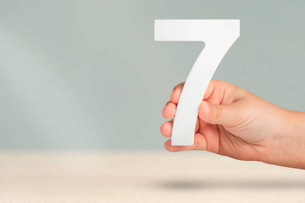 Number seven in hand. Hand holding white number 7 on blurred background with copy space. Concept with number seven. Birthday 7 years, percent, seventh grade or day - Photo, Image