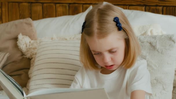 白人の小さな娘が家で本を読んでいる。スマート赤ちゃんの女の子子供で居心地の良いベッドルームで写真を見て教科書で読書文学物語小学校教育で学ぶ - 映像、動画
