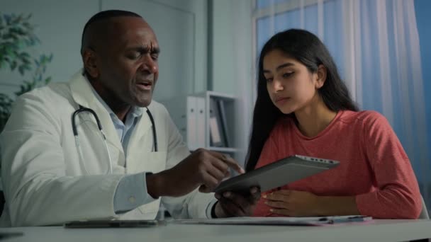 Afrikkalainen keski-ikäinen lääkäri mies lääkäri auttaa nuori nainen potilas tarkistaa sairausvakuutus sähköisen terveystestin tulokset diagnostiikka digitaalinen tabletti näyttää asiakkaan verkossa modernin teknologian lääketieteessä - Materiaali, video