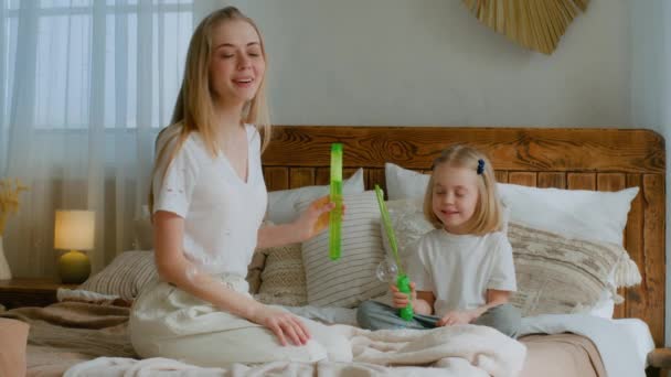 Caucasian mother woman mom with small little child girl kid baby adopted healthy daughter blowing soap bubbles in air in bedroom sitting on bed playing together blow at home family health insurance - Footage, Video