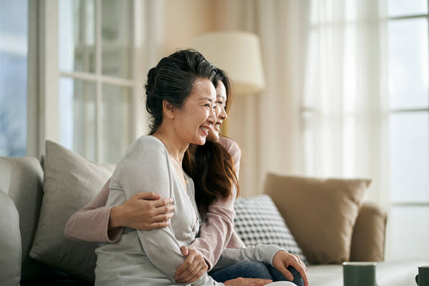 Porträt einer älteren asiatischen Mutter und erwachsenen Tochter, die zu Hause glücklich und lächelnd auf der Couch sitzt - Foto, Bild