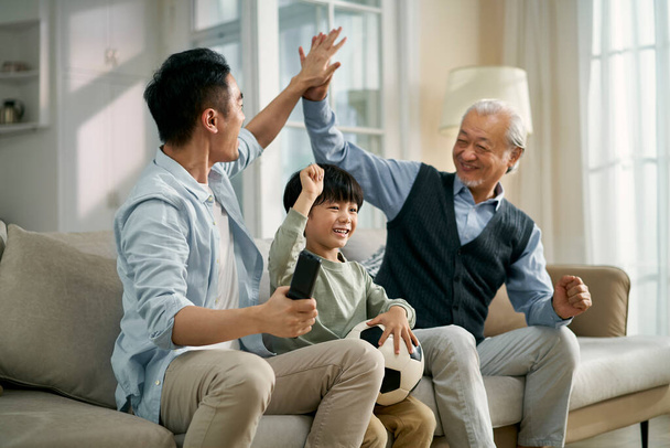 azjatycki syn ojciec dziadek siedzi na kanapie w domu świętując bramkę i zwycięstwo podczas oglądania na żywo transmisji meczu piłki nożnej w telewizji razem - Zdjęcie, obraz