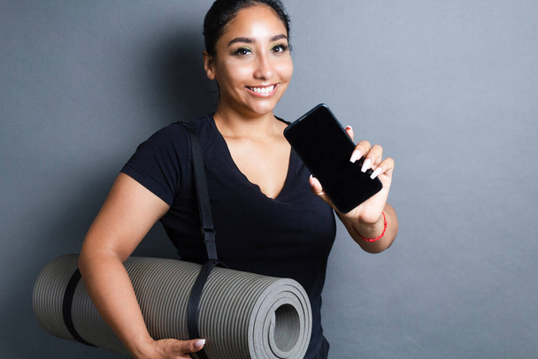 barna hispán fiatal nő bemutató smartphone képernyő, miközben gazdaság jóga mat - Fotó, kép