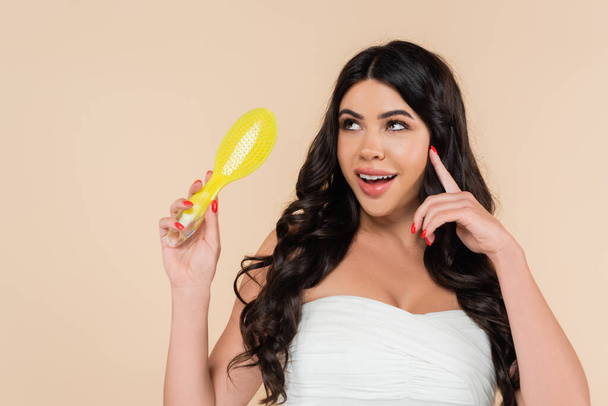Dreamy woman in top holding hairbrush and looking away isolated on beige  - Photo, Image