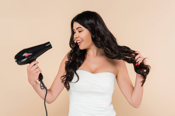 Весела брюнетка жінка тримає фен і торкається волосся ізольовано на бежевому
  - Фото, зображення