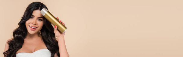 Улыбающаяся брюнетка, держащая лак для волос изолированный на бежевый с копировальным пространством, баннер  - Фото, изображение