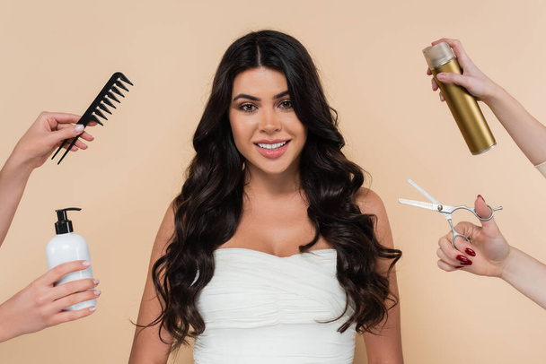 Smiling woman with wavy hair looking at camera near hands with hairdressing tools isolated on beige  - Photo, Image