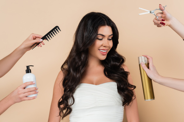 Smiling woman with wavy hairstyle looking at hands holding hairdressing tools isolated on beige  - Photo, Image