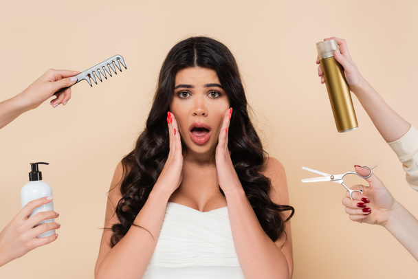 Shocked brunette woman looking at camera near hands with hairdressing tools isolated on beige  - Photo, Image