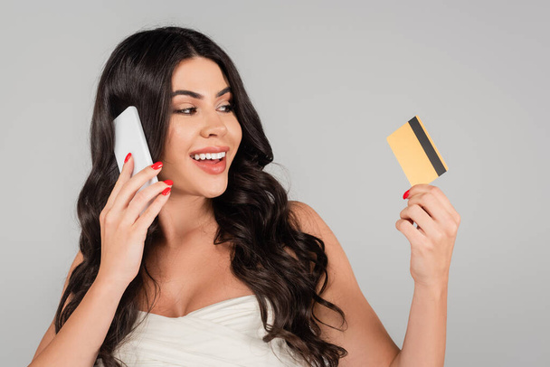 glückliche brünette Frau mit langen Haaren hält Kreditkarte in der Hand und spricht auf dem Smartphone isoliert auf grau - Foto, Bild