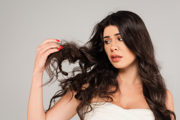 displeased brunette woman looking at long and tousled hair isolated on grey - Foto, imagen