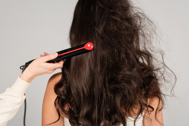 hairdresser with hair iron straightening wavy and tousled hair of brunette woman isolated on grey - Photo, image