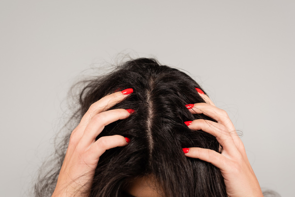 oříznutý pohled na brunetku s červenou manikúrou dotýkající se hlavy s rozcuchanými vlasy izolované na šedé - Fotografie, Obrázek