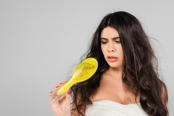 sad brunette woman with tangled hair looking at hair brush isolated on grey - Photo, image