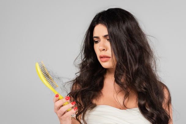 traurige brünette Frau schaut auf Haarbürste mit verlorenen Haaren isoliert auf grau - Foto, Bild