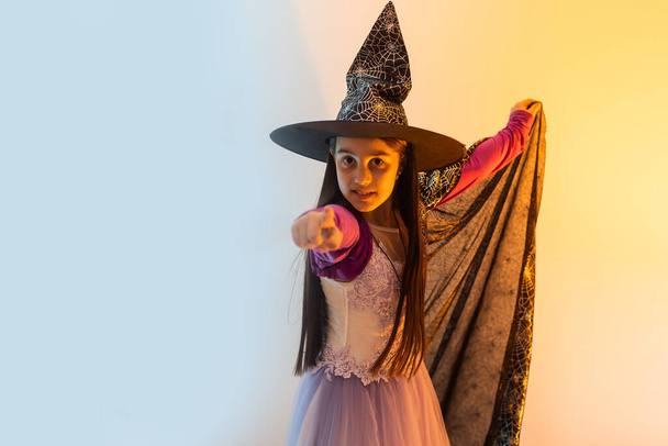 Happy scary Halloween concept: little witch girl playing paranormal Halloween role and doing scary look on a yellow background. Kid scaring and making Boo in carnival witch costume or wizard costume. - Photo, Image