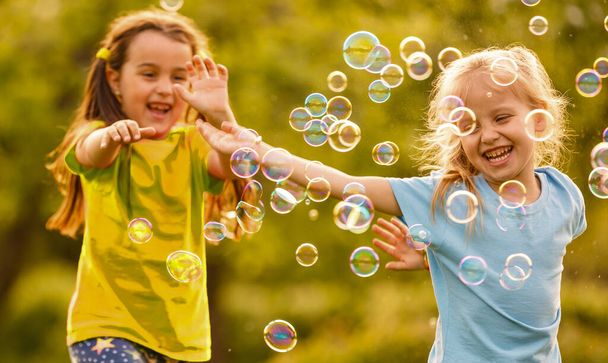 Two cheerful cute girls are playing outdoors with huge soap bubbles. - Photo, Image
