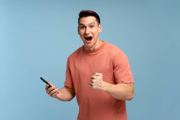 Šťastný spokojený muž v tričku drží smartphone a usmívá se, takže ano gesto, slaví on-line loterie nebo rozdávat vítězství. Vnitřní studio záběr izolované na modrém pozadí  - Fotografie, Obrázek