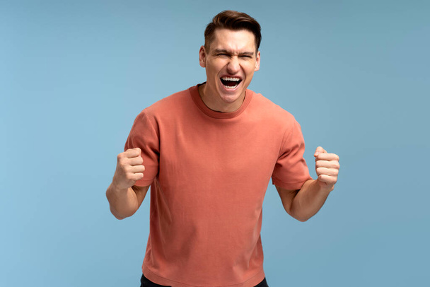 Portrait of happy screaming handsome young man standing with suprised face and rejoicing his victory. Indoor studio shot, isolated on blue background  - Photo, Image
