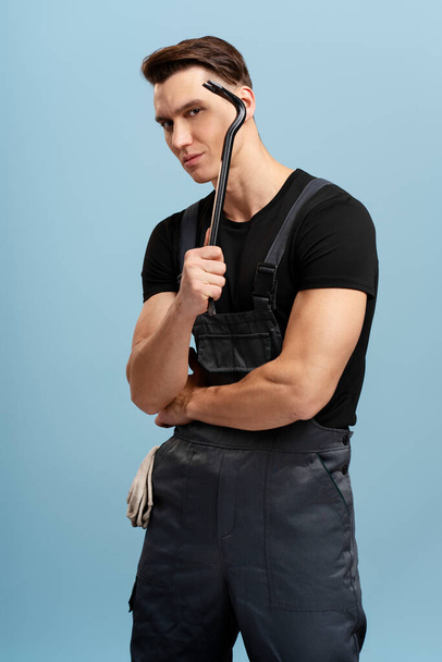 Serious male builder holding crowbar isolated on blue background. Man in uniform with claw hammer in his hand, selective focus  - Photo, Image