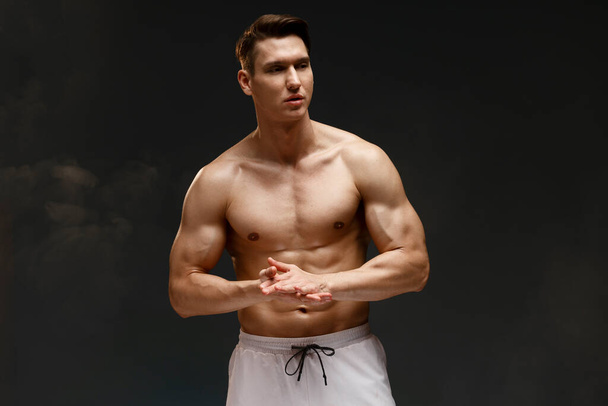 Homem fisiculturista caucasiano grave com tronco muscular nu vestindo shorts posando isolado no fundo escuro  - Foto, Imagem