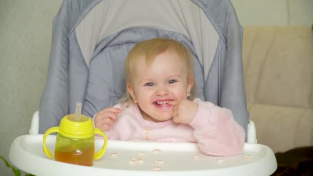 Een lief anderhalf jaar oud meisje zit in een hoge babyvoedingsstoel en eet koekjes. - Video