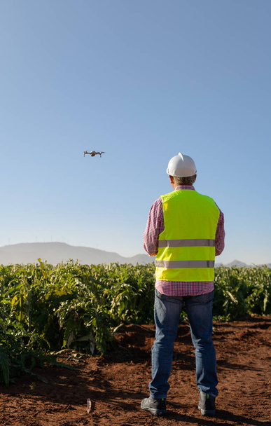Agronomist with a vest and helmet piloting a drone over a sustainable crop field. Experienced older man. in the background some mountains and a wind farm. Technology concept for agronomy industry, ecology. vertical photo. - Photo, Image