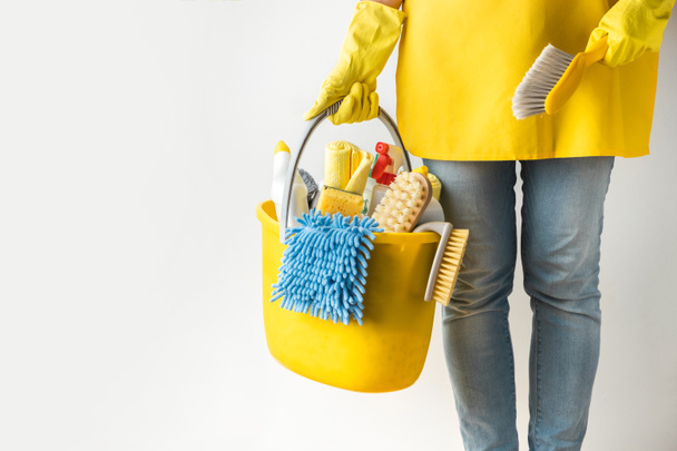 Unrecognizable young caucasian woman in a uniform wearing yellow rubber gloves and holding bucket full of cleaning supplies. Cleaning company service advertisement. House cleaning concept - Photo, image