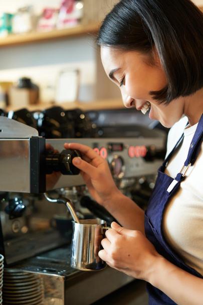 Szczęśliwa Azjatka, baristka używająca ekspresu do kawy do zamawiania, parujące mleko do cappuccino i latte, śmiejąca się i uśmiechnięta podczas pracy w kawiarni. - Zdjęcie, obraz