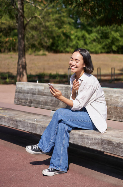 スタイリッシュな若いアジアの女の子、スマートフォンで公園に座っている学生、電話を使用して、ベンチに座っている間誰かを待っています. - 写真・画像