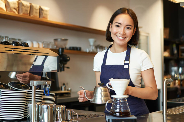 Kahveci kız, Asyalı barmen çaydanlıktan su dolduruyor, tezgahın arkasındaki kafede filtre kahve yapıyor, mavi önlük giyiyor.. - Fotoğraf, Görsel
