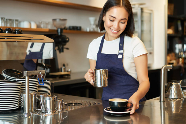 Krásná mladá žena barista, dělat cappuccino, nalévání vařené mléko pro latte art do poháru, dát svou objednávku za pultem. - Fotografie, Obrázek